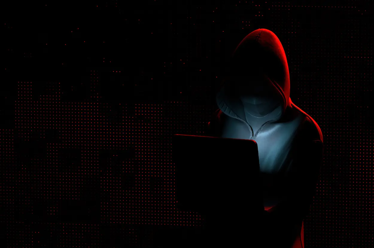 Hooded figure lit by a laptop in a dark room.