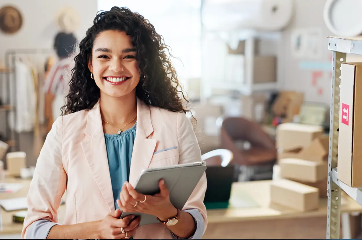 Smiling woman in a pastel blazer holding a tablet in a modern workspace with boxes and clothing rack in the background.