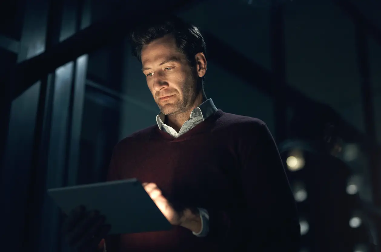 Man in a dark room focused on a tablet, showcasing modern technology and professional engagement.