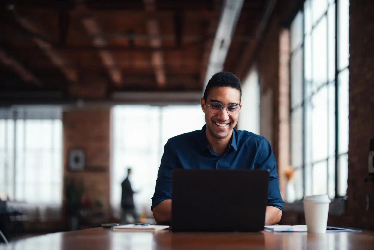 Smiling man working on a laptop in a modern office with large windows and a coffee cup on the table.