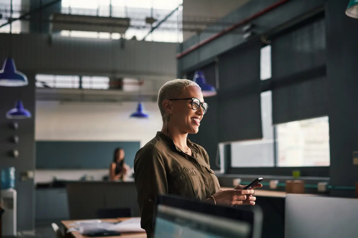 Smiling woman in glasses holding a smartphone in an office.