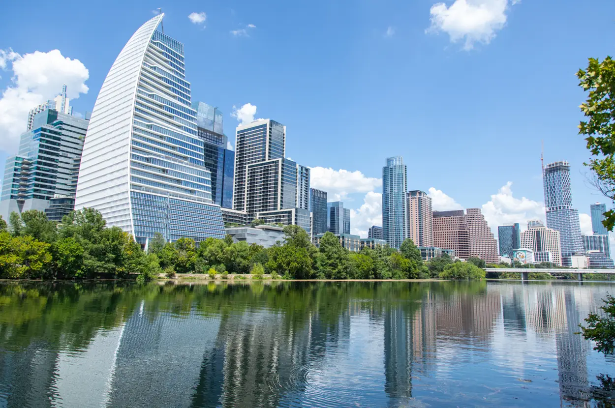 Austin skyline reflecting on water with modern architecture and lush green trees under a bright blue sky.