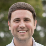 Smiling man in a casual shirt standing outdoors with greenery in the background.