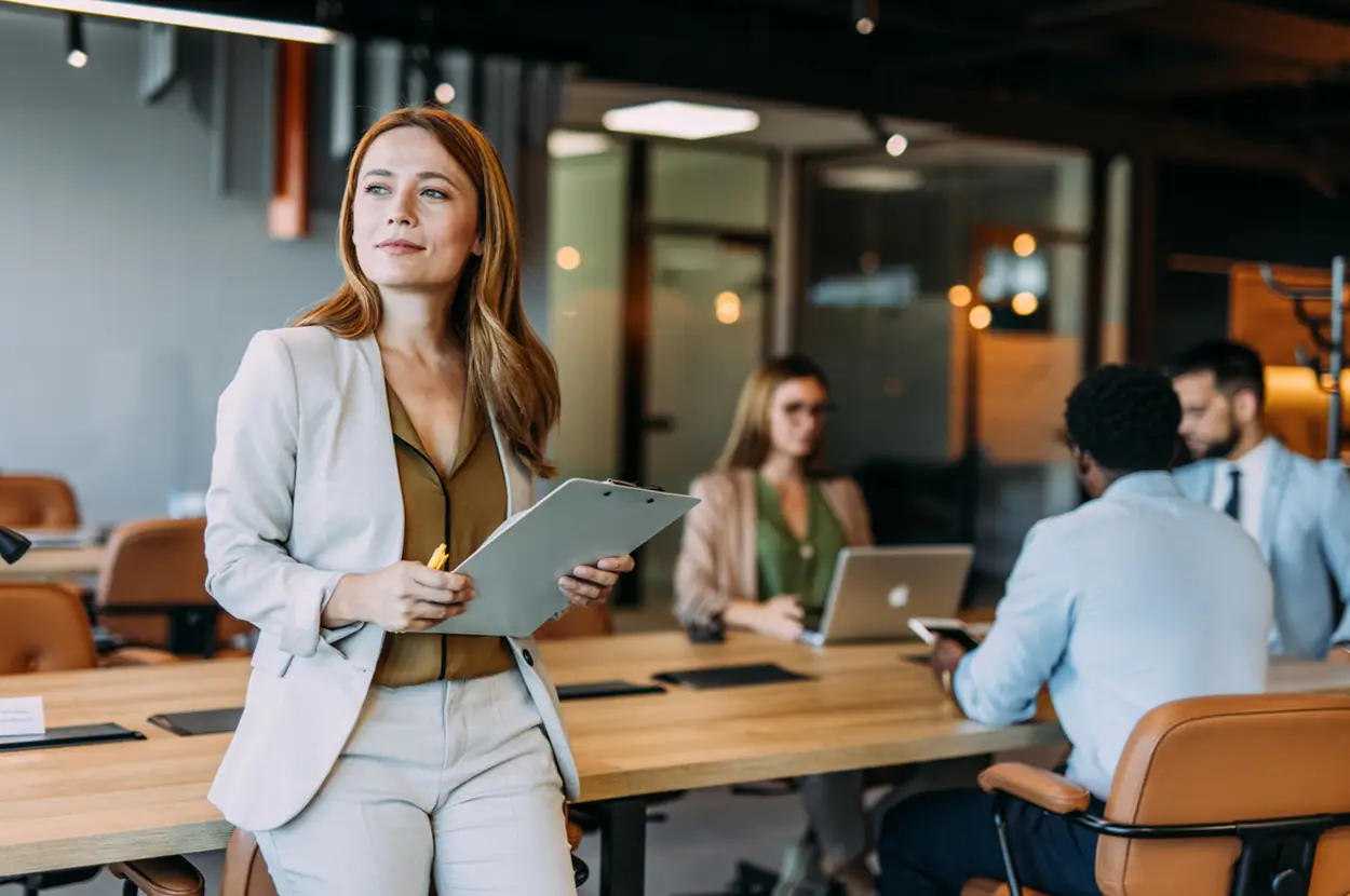 Businesswoman in a modern office meeting, holding a clipboard, with colleagues working in the background.