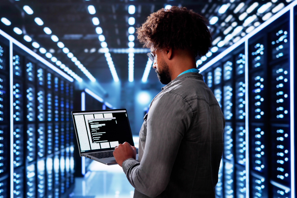 A professional analyzing data on a laptop in a modern server room with blue lighting and server racks.