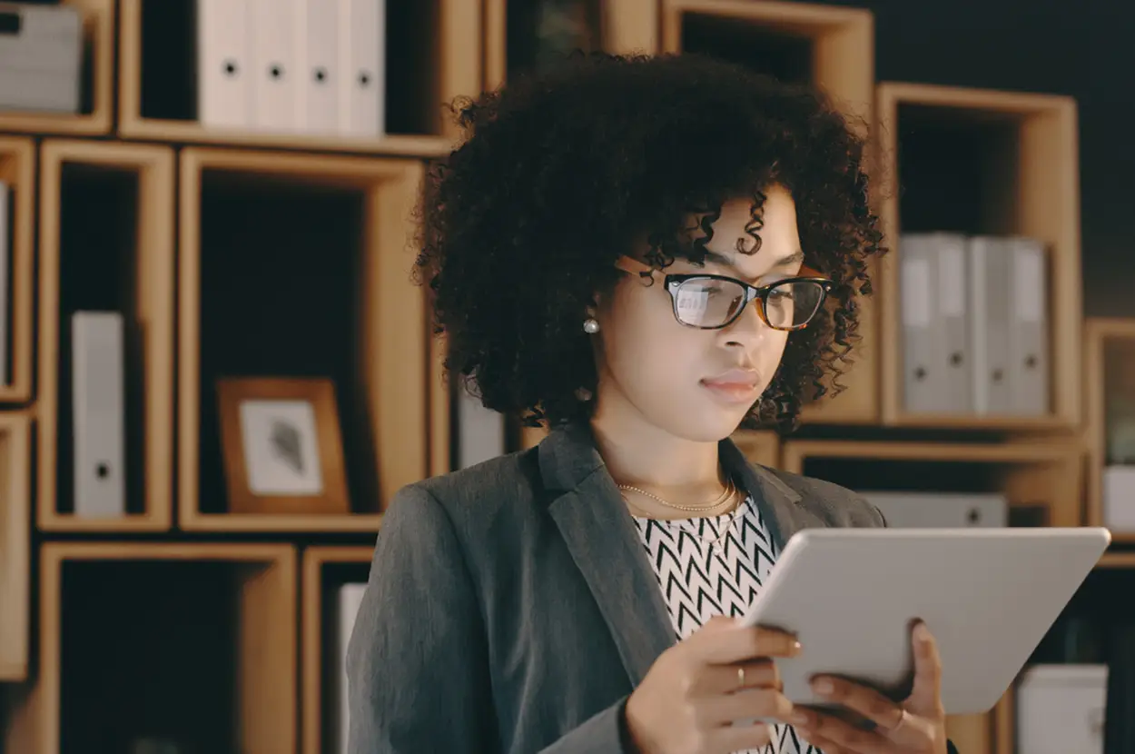 Professional woman with glasses using a tablet in a modern office, with shelves of files in the background.
