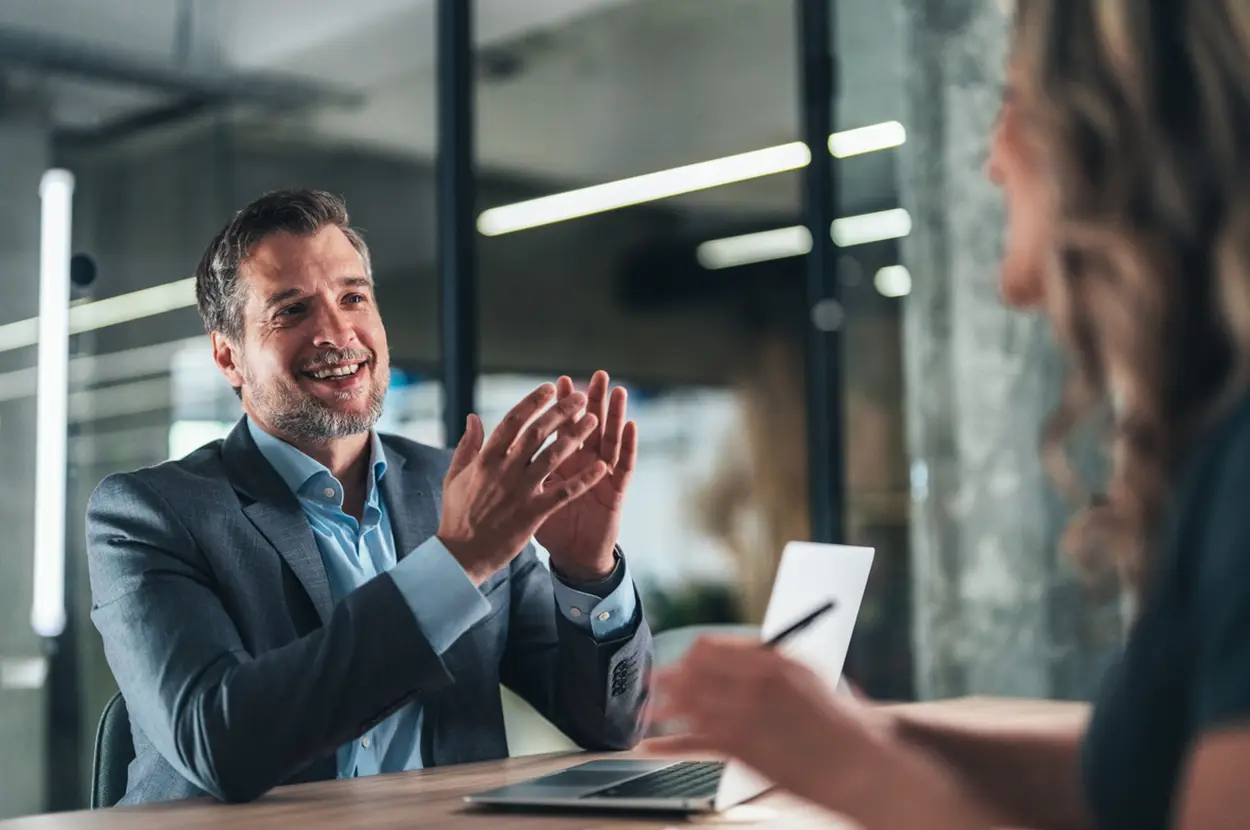 Business meeting in a modern office, featuring a smiling man engaging with a woman taking notes on a laptop.