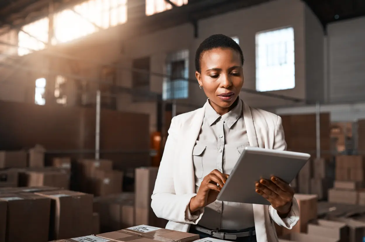 Woman in a warehouse using a tablet to manage inventory, surrounded by stacked boxes in a well-lit environment.