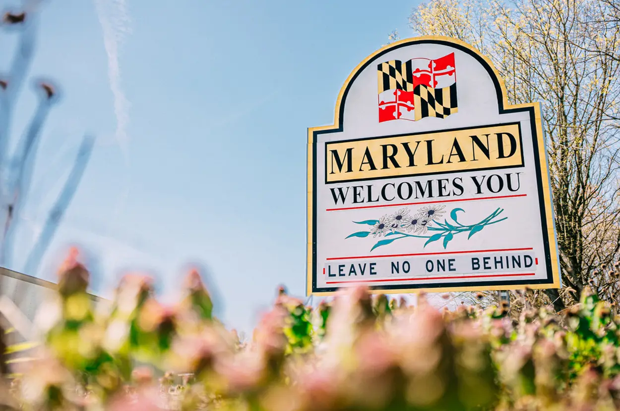Welcome sign for Maryland featuring its state flag and the phrase 'Leave No One Behind' against a clear blue sky.