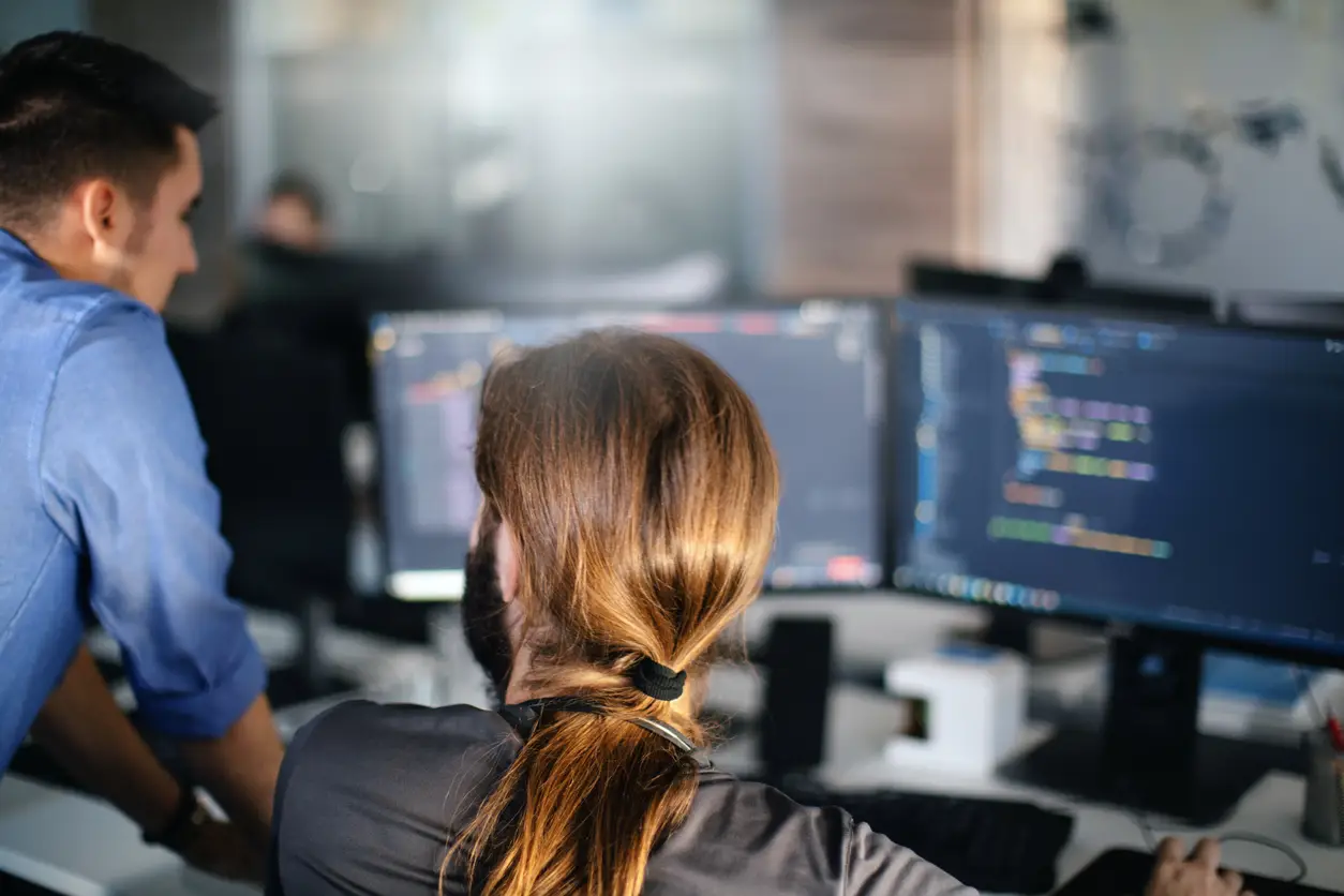 Two programmers collaborate at a computer, analyzing telemetry on dual screens in a modern office setting.