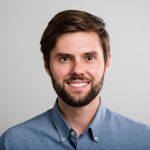 Smiling young man in a blue shirt against a light gray background, showcasing a friendly and approachable demeanor.