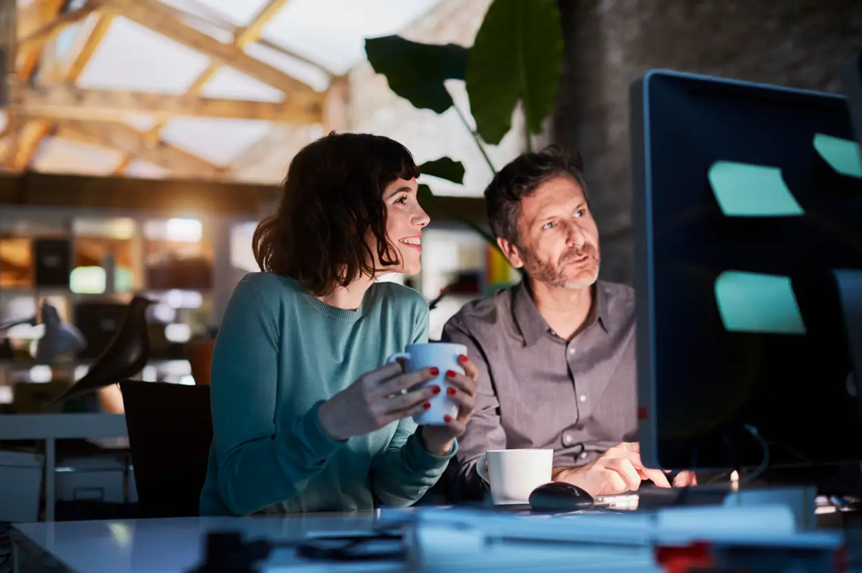 Two colleagues discussing ideas at a computer in a modern office setting, surrounded by plants and creative decor.