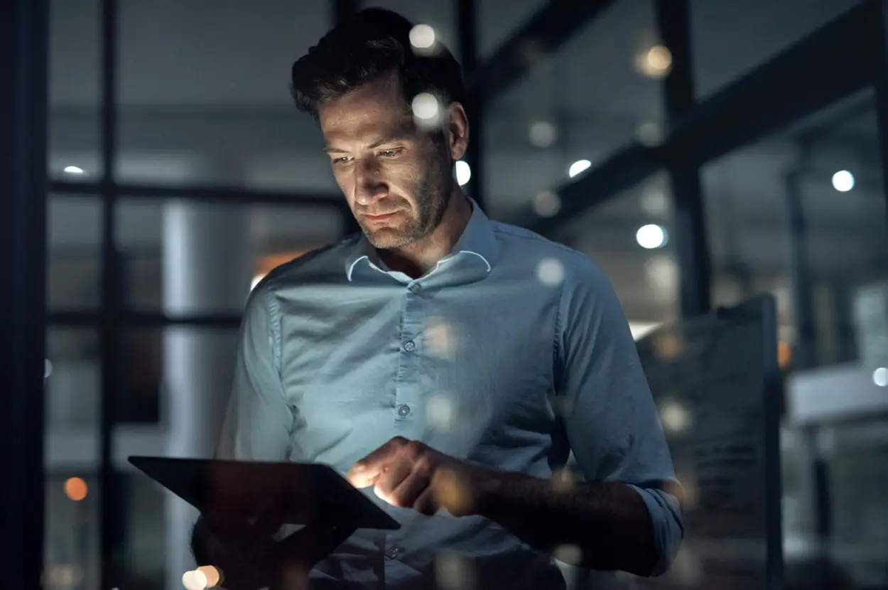 Man using a tablet in a modern office at night, focused on work with city lights in the background.