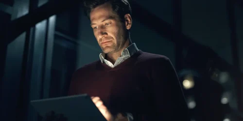 Man in a dark room focused on a tablet, showcasing modern technology and professional engagement.