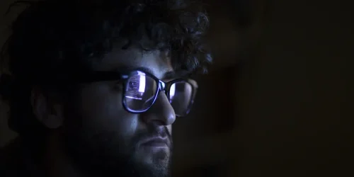 Person with curly hair and glasses focused on a screen, illuminated by soft light in a dark room.