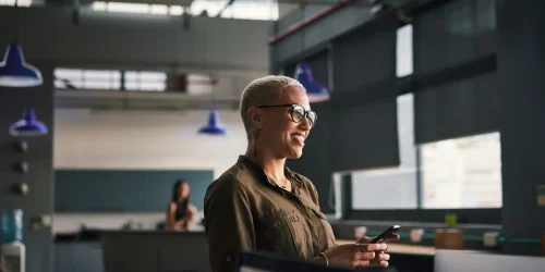 A smiling woman in glasses holding a smartphone in a modern office setting, with a blurred background of coworkers.