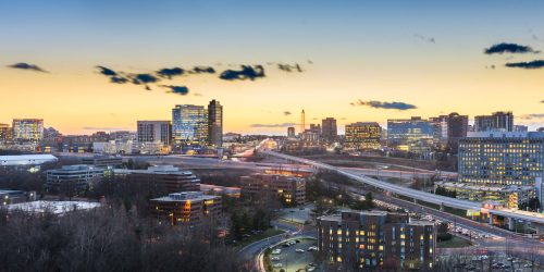 Panoramic view of a city skyline at dusk, featuring modern buildings and highways under a colorful sunset.