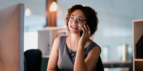 Woman in a gray dress smiles while on the phone in a modern office setting with a computer and stylish decor.