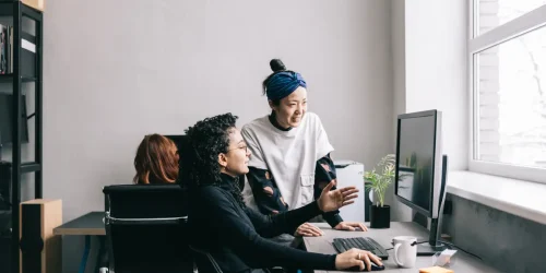 Two women collaborate in a modern workspace, discussing ideas while working on a computer, enhancing teamwork.