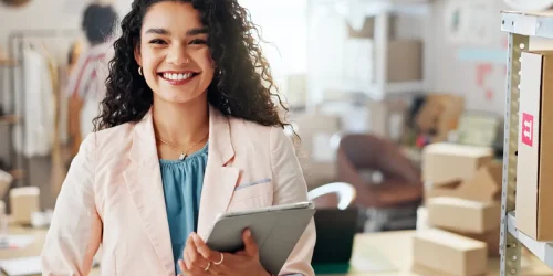 Smiling woman in a pastel blazer holding a tablet in a modern workspace with boxes and clothing rack in the background.