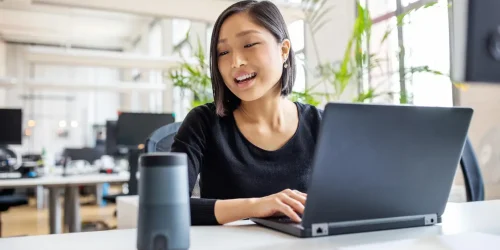 Young woman working on a laptop in a modern office, smiling with a smart speaker on the table nearby.